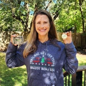 Woman in a "Trail Racing Over Texas — Brazos Bend 100" hoodie holding up two race belt buckles in a backyard