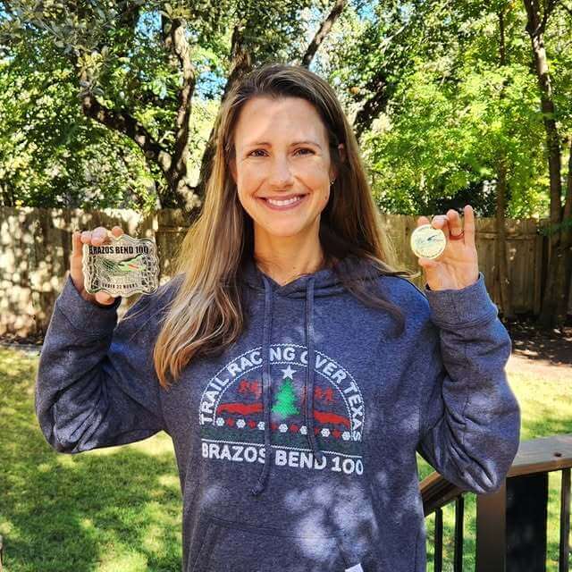 Woman in a "Trail Racing Over Texas — Brazos Bend 100" hoodie holding up two race belt buckles in a backyard