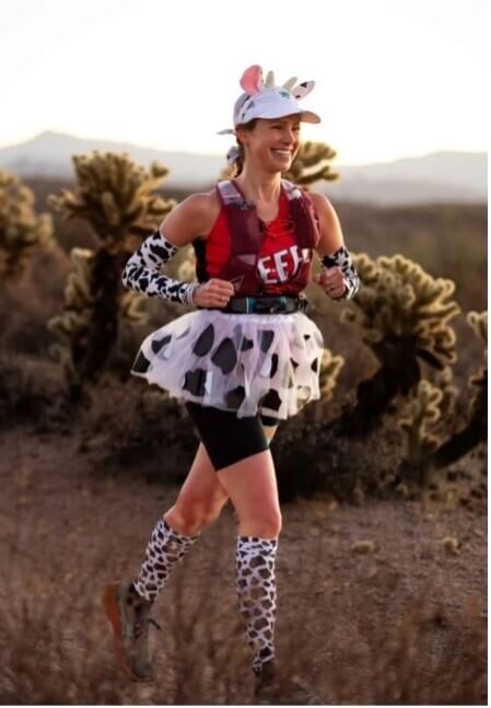 Woman trail running in a cow-print costume with ears, a tutu, and spotted leg warmers through a desert landscape with cacti